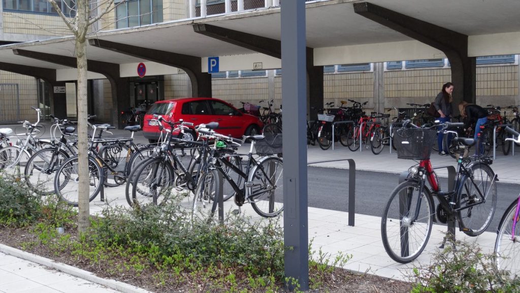 A row of parked bicycles lined up neatly along a sidewalk in a urban setting.